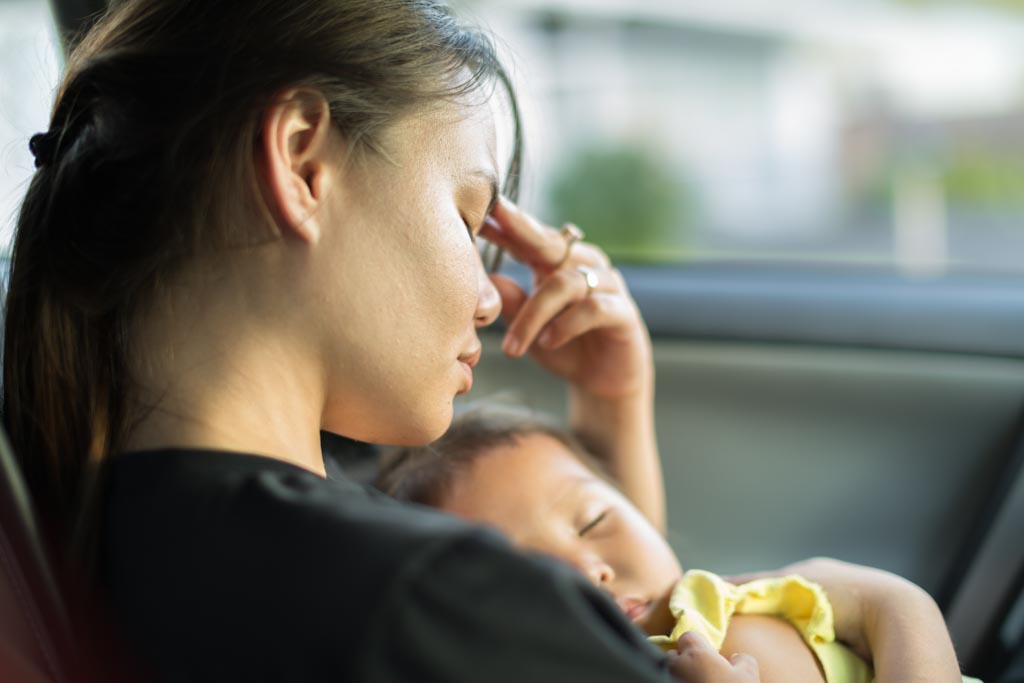 Mother holding her sleeping baby with her eyes closed and her hand resting on her forehead.